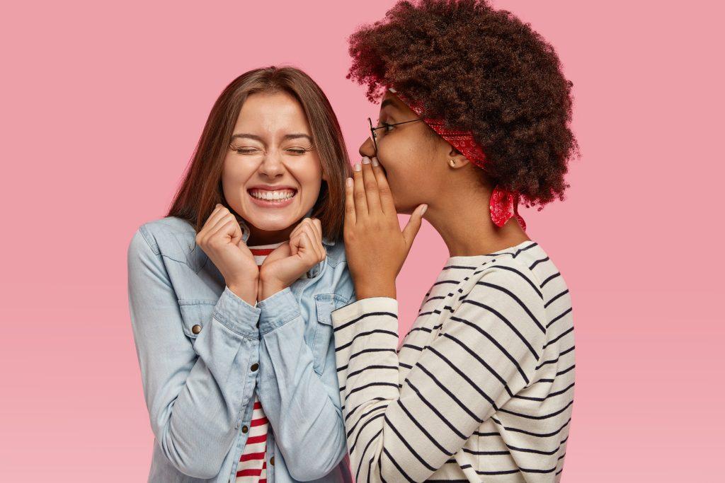 black ethnic woman whispers secret to her caucasian female with toothy smile gossip together scaled